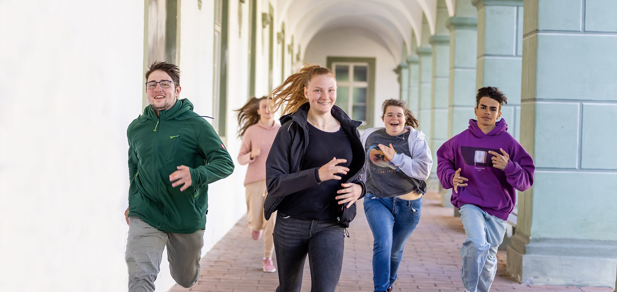 Fünf lachende Teenager rennen fröhlich durch einen historischen Bogengang mit Säulen und Bögen.