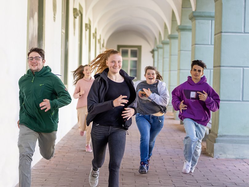 Fünf lachende Teenager rennen fröhlich durch einen historischen Bogengang mit Säulen und Bögen.