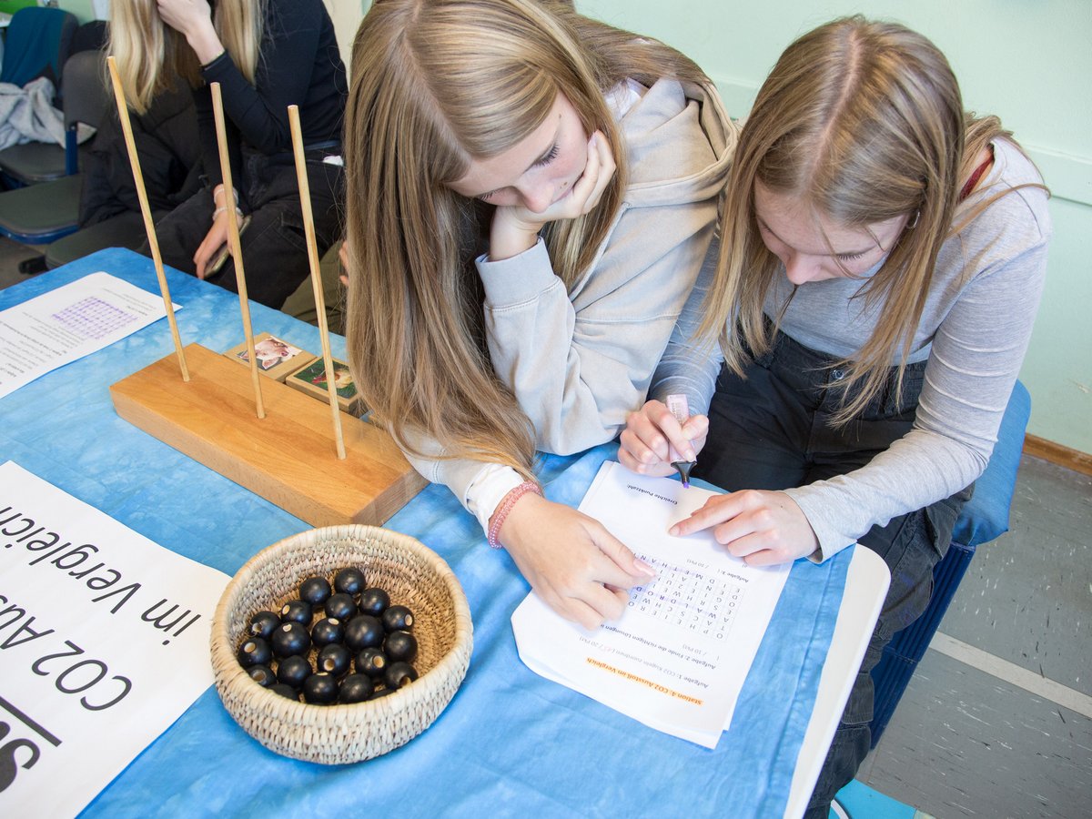Zwei Frauen lösen ein Rätsel am Tisch mit Schild 'CO2-Ausstoß im Vergleich', einem Korb mit Perlen und Holzspiel.