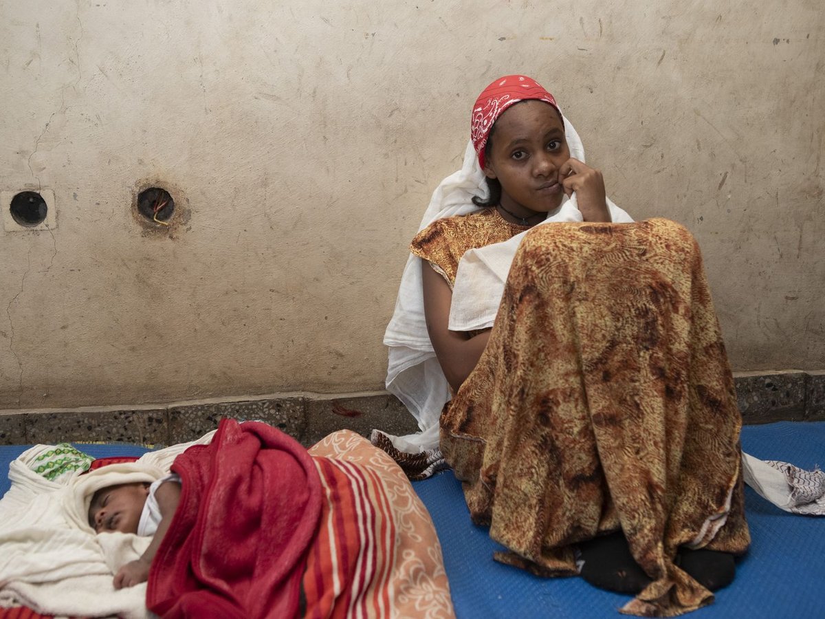 Eine Frau mit rotem Kopftuch sitzt nachdenklich auf einer Matte neben einem schlafenden Baby vor einer kahlen Wand mit freiliegenden Kabeln.