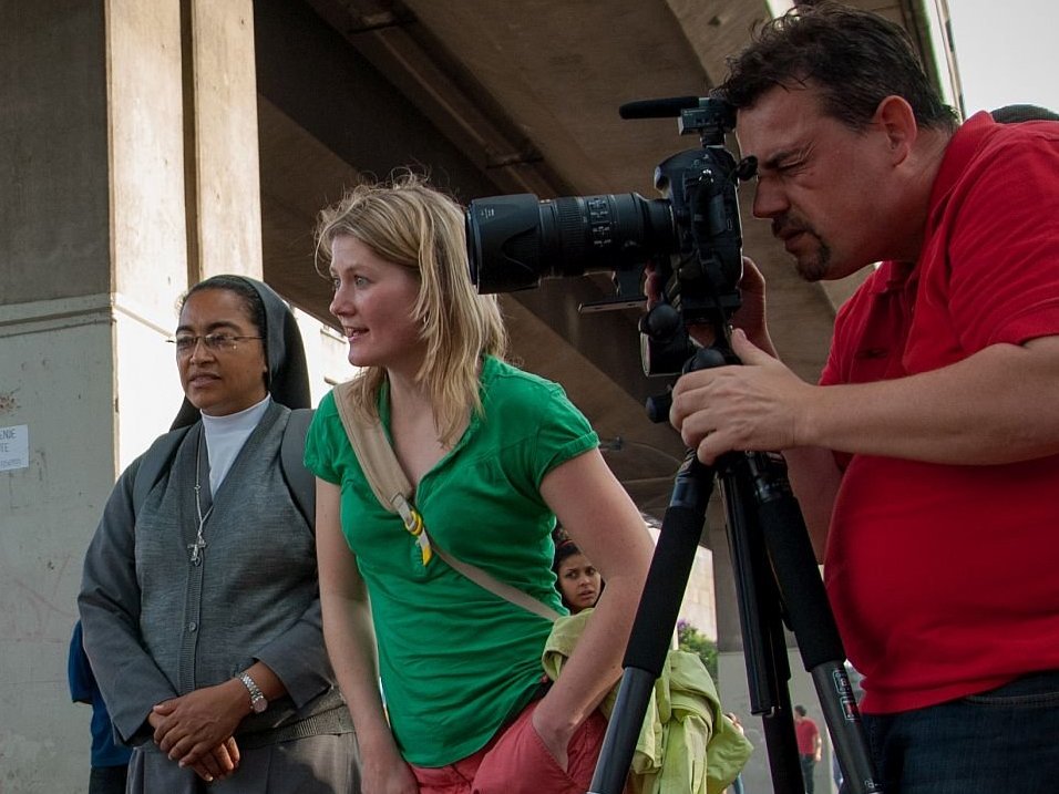 Ein Kameramann filmt eine junge Frau auf einer belebten Straße, beobachtet von einer Nonne und einer anderen Frau.