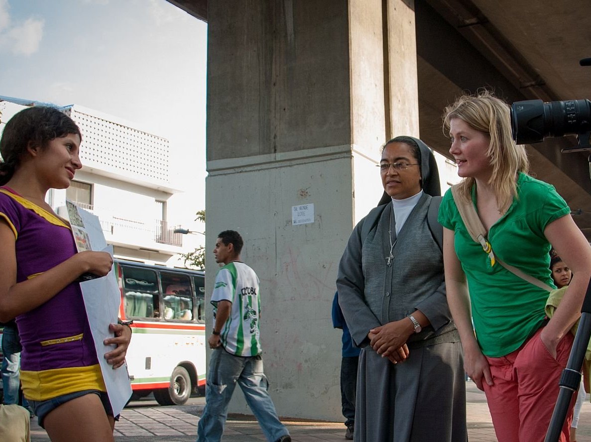 Ein Kameramann filmt eine junge Frau auf einer belebten Straße, beobachtet von einer Nonne und einer anderen Frau.