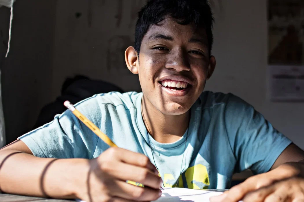 Ein lächelnder junger Mensch mit dunklem Haar schreibt mit einem Bleistift an einem sonnigen Schreibtisch.