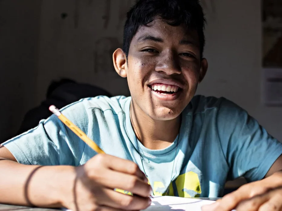 Ein lächelnder junger Mensch mit dunklem Haar schreibt mit einem Bleistift an einem sonnigen Schreibtisch.