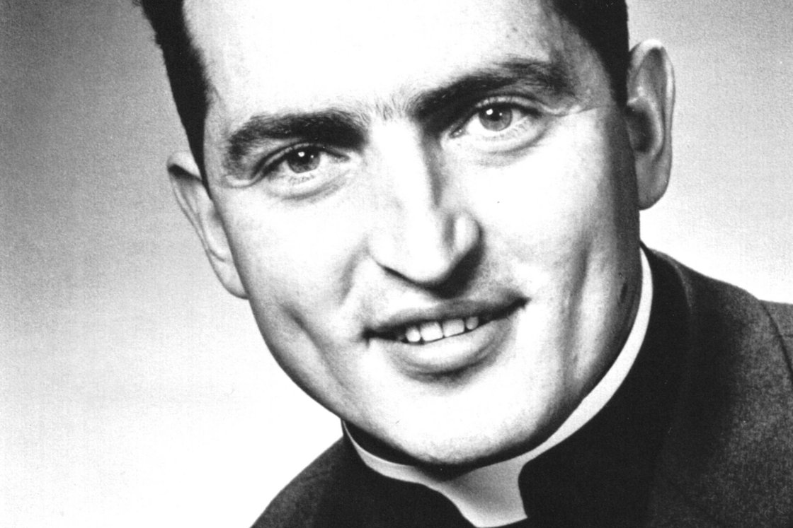 Schwarz-Weiß-Porträt eines lächelnden jungen Priesters mit dunklem Haar und Priesterkragen, direkt in die Kamera blickend.