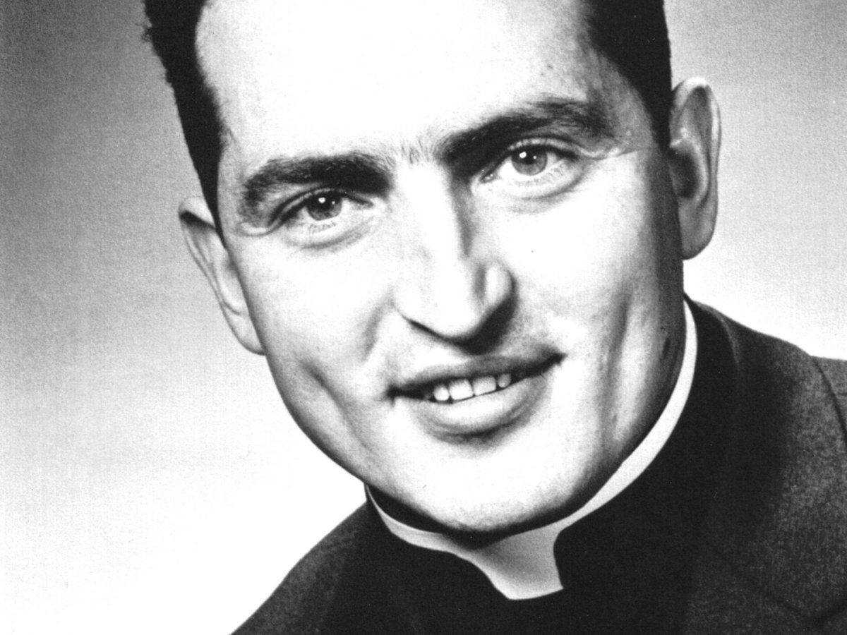 Schwarz-Weiß-Porträt eines lächelnden jungen Priesters mit dunklem Haar und Priesterkragen, direkt in die Kamera blickend.