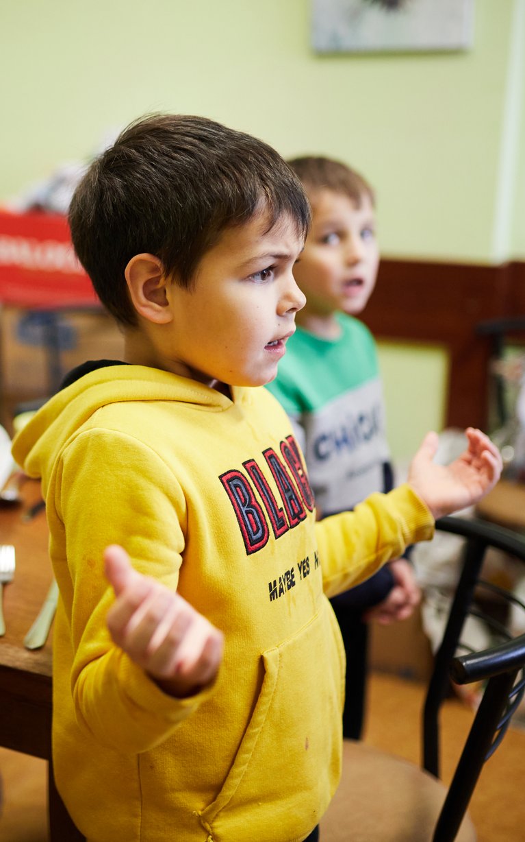 Drei kleine Jungen, darunter einer im gelben Kapuzenpullover, gestikulieren an einem Tisch mit Kanne und Brot.