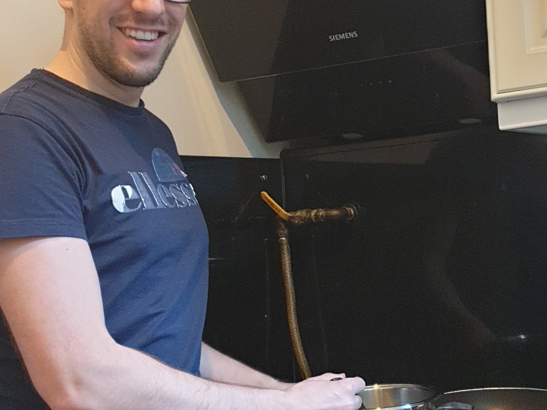 Ein lächelnder Mann mit Brille und Schürze kocht auf einem Gasherd und hält einen Topf über einer blauen Flamme neben einer Pfanne.