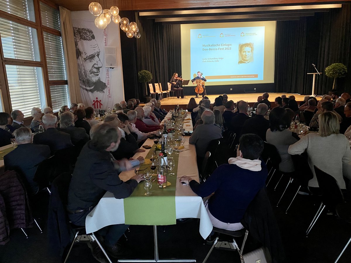 Zuschauer beim Don-Bosco-Fest sehen zwei Musiker (Geige, Cello) auf einer Bühne. Ein Bildschirm zeigt Veranstaltungsinformationen.