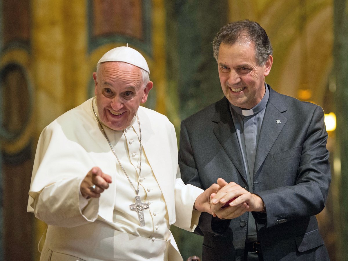 Papst Franziskus in Weiß lächelt fröhlich und zeigt, während er Hände mit einem lächelnden Priester hält.