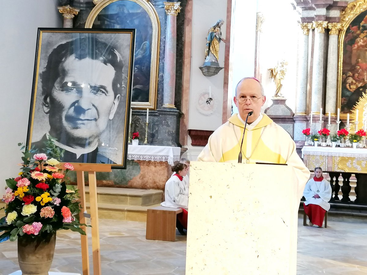 Ein Priester hält eine Predigt in einer Kirche, flankiert von einem Porträt einer religiösen Figur und einem Blumenschmuck.