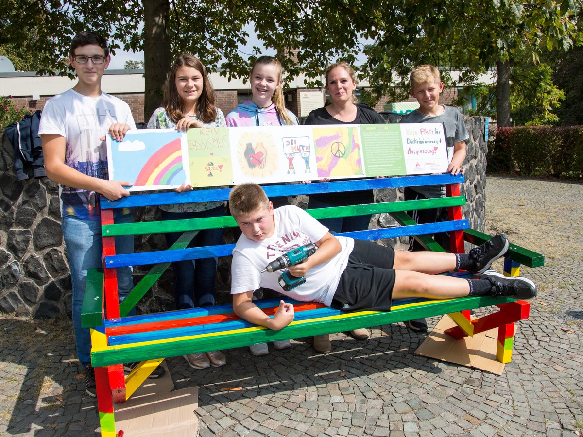 Schüler und ein Erwachsener posieren mit einer regenbogenfarbenen Anti-Rassismus-Bank und Schildern. Ein Junge liegt mit einem Bohrer darauf.