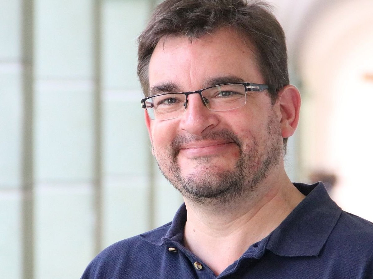 Ein lächelnder Mann mit Brille und Bart in einem blauen Poloshirt. Im Hintergrund ist ein verschwommenes Kruzifix zu sehen.