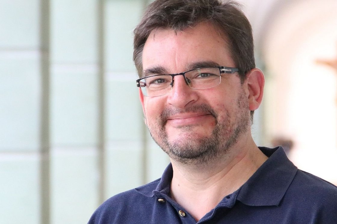 Ein lächelnder Mann mit Brille und Bart in einem blauen Poloshirt. Im Hintergrund ist ein verschwommenes Kruzifix zu sehen.