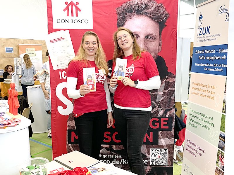 Zwei lächelnde Frauen in roten Don Bosco T-Shirts stehen an einem Messestand und halten Broschüren.