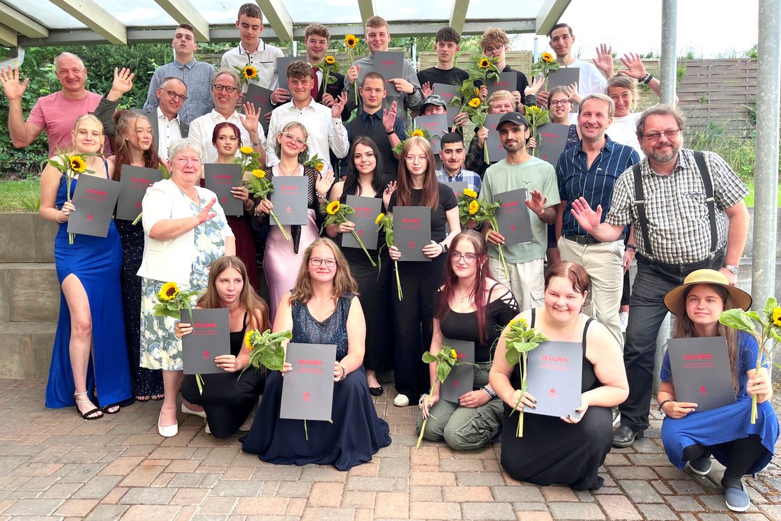 Große Gruppe von Schülern und Erwachsenen, viele halten 'ZEUGNIS'-Zertifikate und Sonnenblumen, lächeln und winken bei einer Zeremonie im Freien.