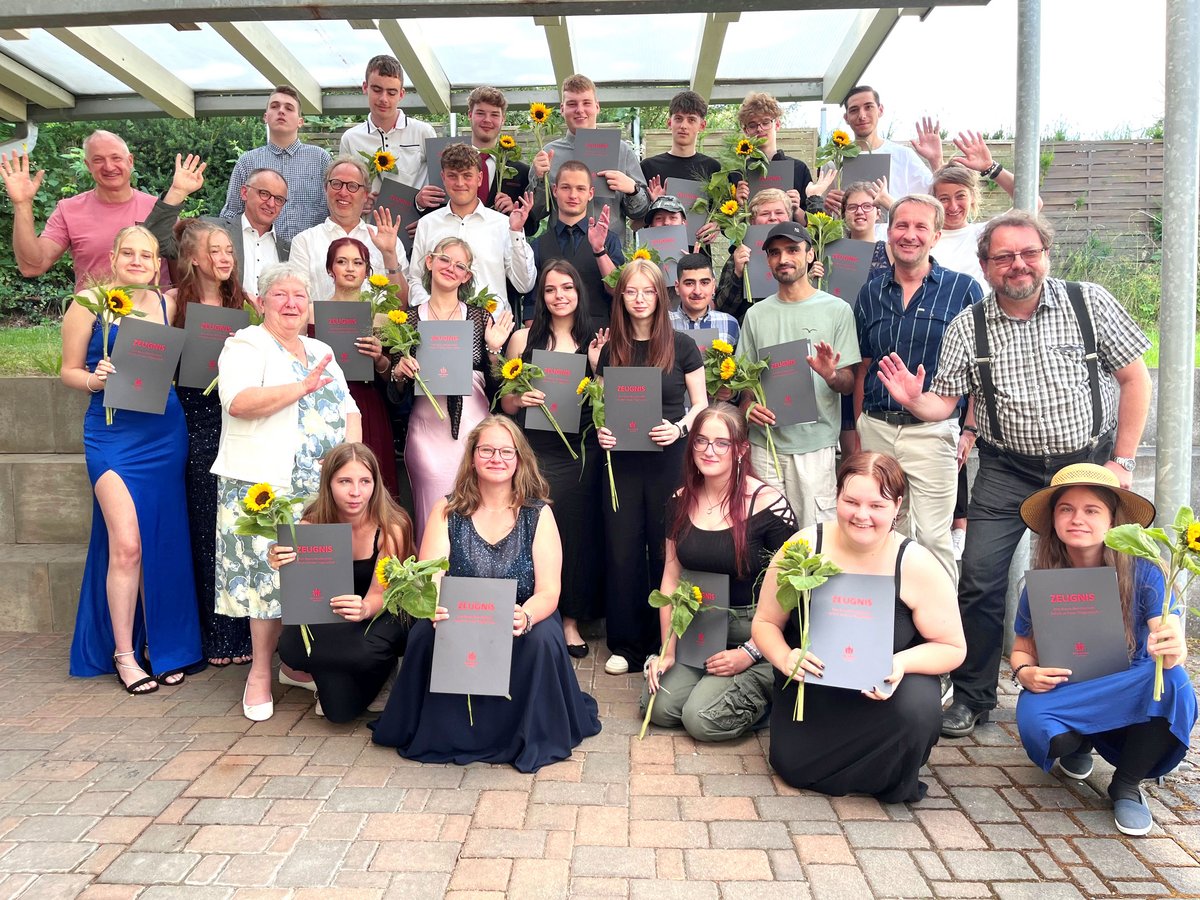 Große Gruppe von Schülern und Erwachsenen, viele halten 'ZEUGNIS'-Zertifikate und Sonnenblumen, lächeln und winken bei einer Zeremonie im Freien.