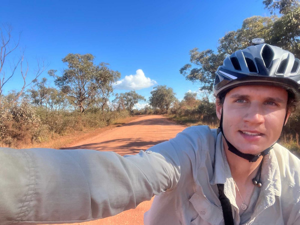 Selfie eines Radfahrers mit Helm auf einer roten Erdstraße, gesäumt von Bäumen unter blauem Himmel.