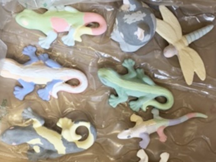 Handbemalte Keramikfiguren wie Eidechsen und eine Libelle, in Pastellfarben auf einer Plastikfolie liegend.