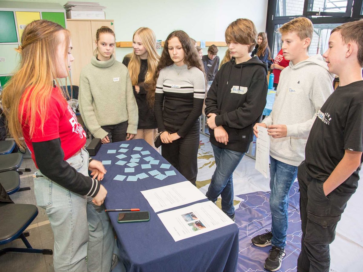 Eine Gruppe jugendlicher Schüler versammelt sich um einen Tisch mit blauen Karten und Dokumenten und lauscht einem Mädchen in Rot.