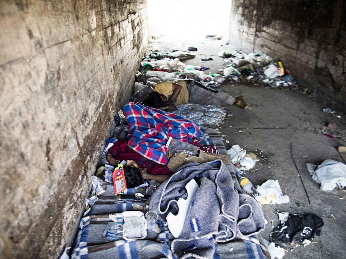 Obdachlose schlafen auf Decken in einem schmutzigen, vermüllten Betontunnel, der Armut und Vernachlässigung zeigt.