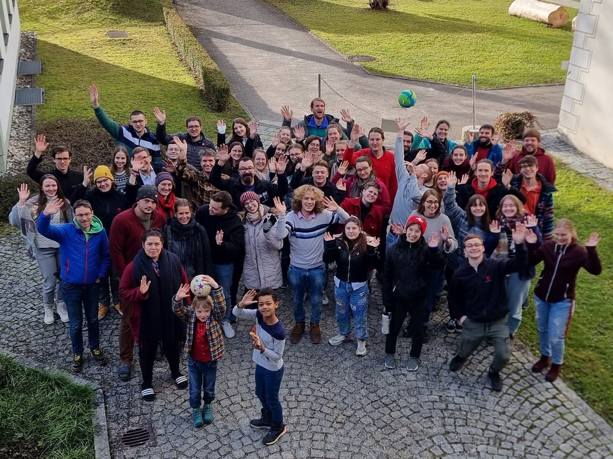Eine vielfältige Gruppe Menschen jeden Alters, inklusive Kindern, winkt fröhlich in die Kamera auf einem Kopfsteinpflasterweg.