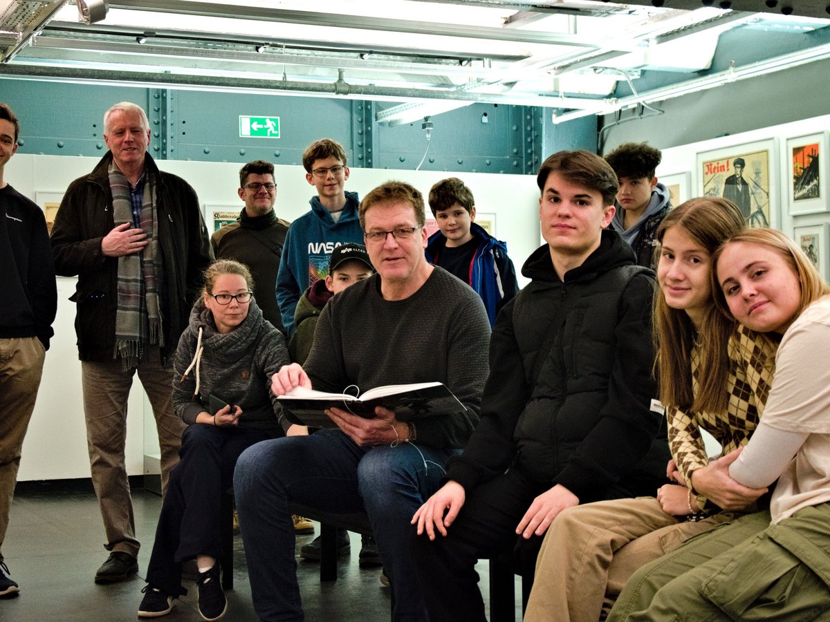 Eine Gruppe von Jugendlichen und Erwachsenen in einer Ausstellung. Ein Mann sitzt mit einem Buch, andere stehen oder sitzen um ihn herum.