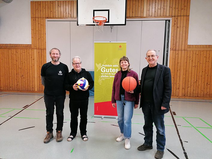 Vier lächelnde Personen in einer Turnhalle, zwei halten einen Volleyball und Basketball, vor einem 'Gutes tun'-Banner.