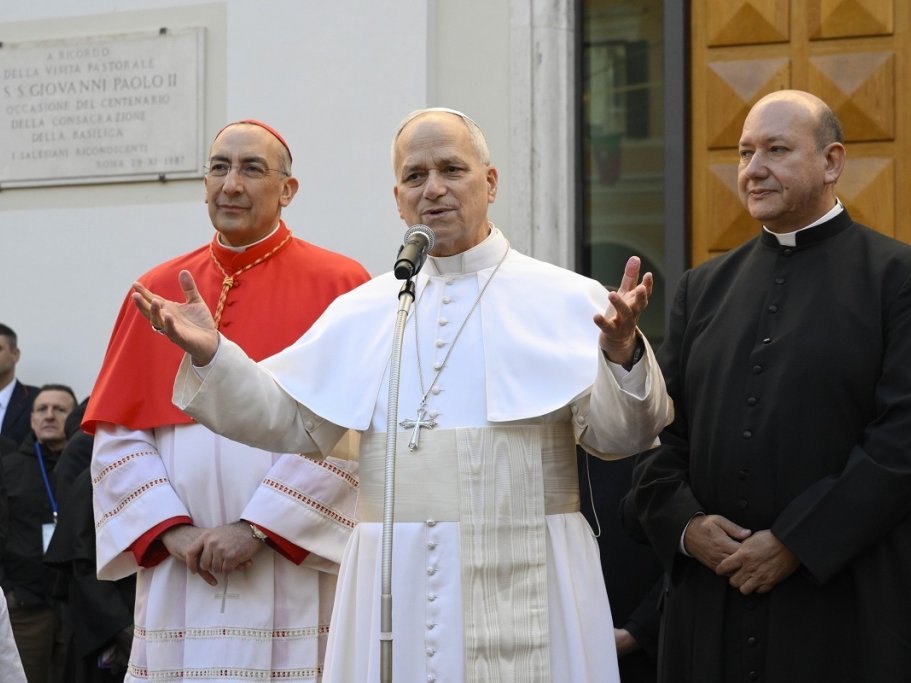 Ein Pontifex in Weiß spricht ins Mikrofon, flankiert von einem Kardinal in Rot und einem Priester in Schwarz.
