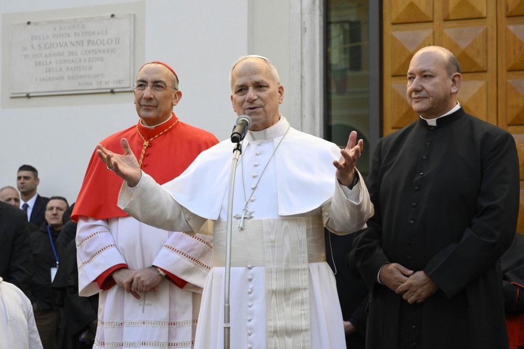 Ein Pontifex in Weiß spricht ins Mikrofon, flankiert von einem Kardinal in Rot und einem Priester in Schwarz.