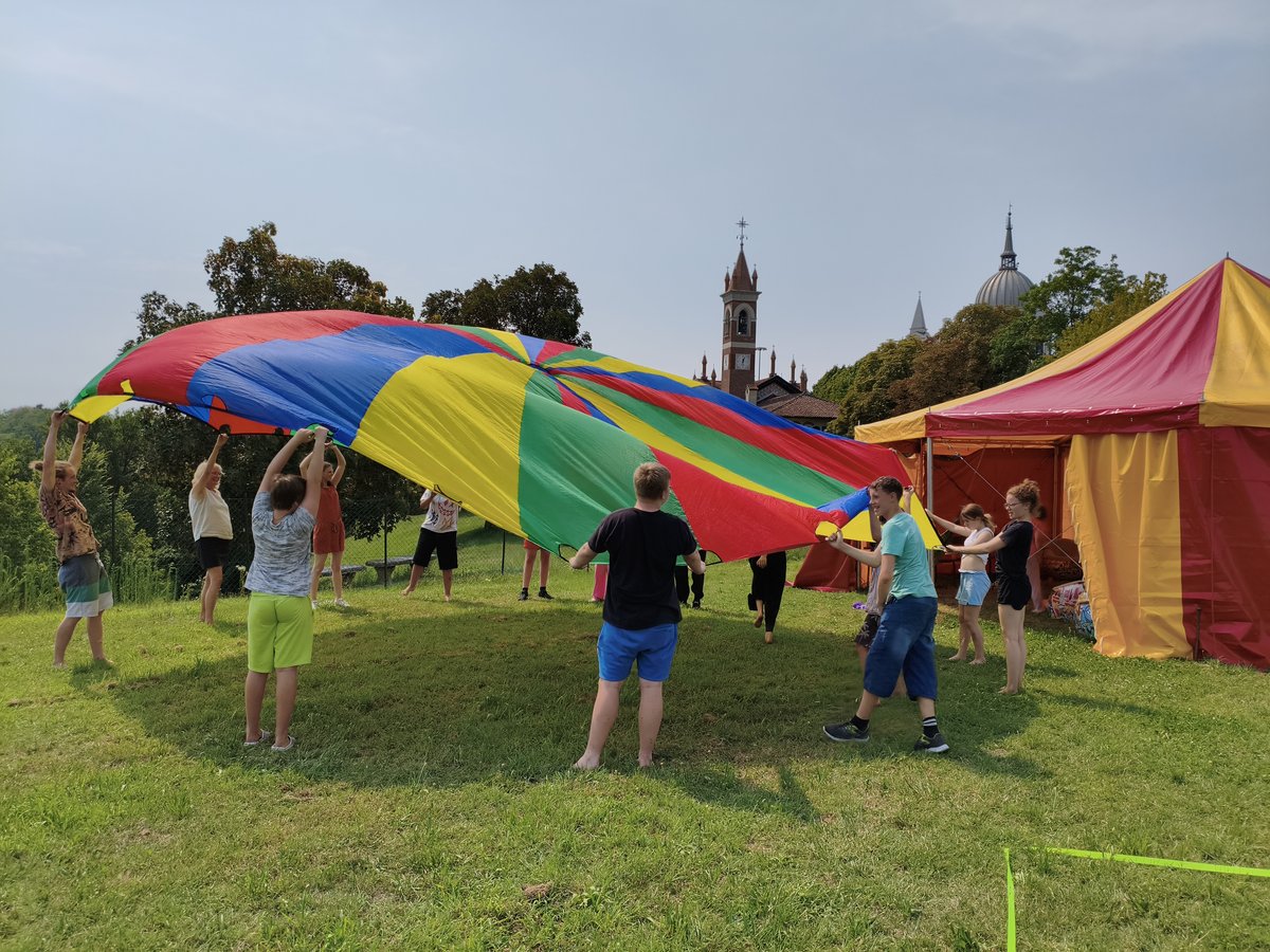 Menschen verschiedenen Alters spielen mit einem großen Regenbogen-Fallschirm auf einer sonnigen Wiese, mit Zelten und Kirchtürmen im Hintergrund.