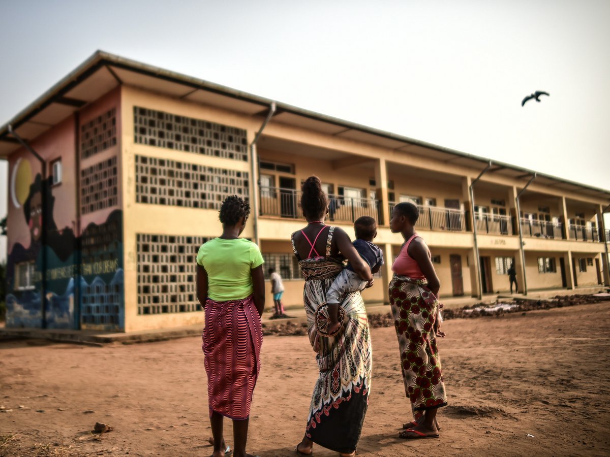 Drei Frauen und ein Kind stehen auf schmutzigem Boden, mit dem Rücken zur Kamera, vor einem Gebäude mit Wandgemälde. Ein Vogel fliegt.
