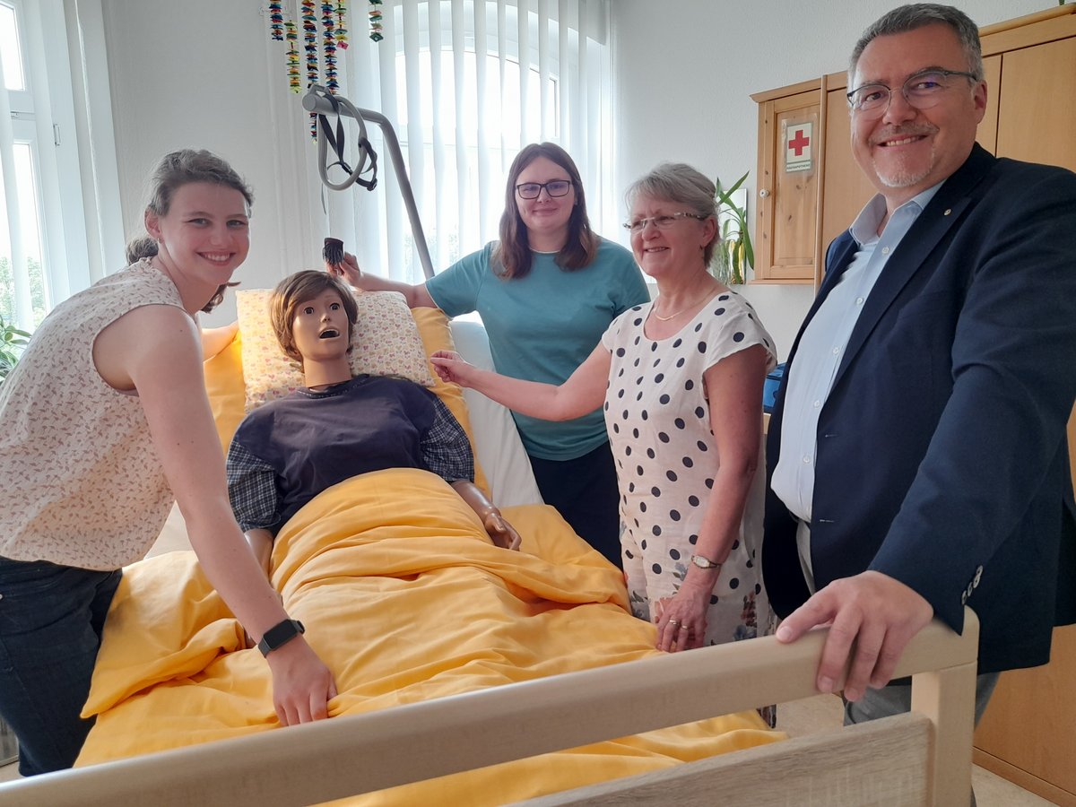 Vier lächelnde Personen, darunter drei Frauen und ein Mann, stehen um eine medizinische Übungspuppe in einem Krankenhausbett.