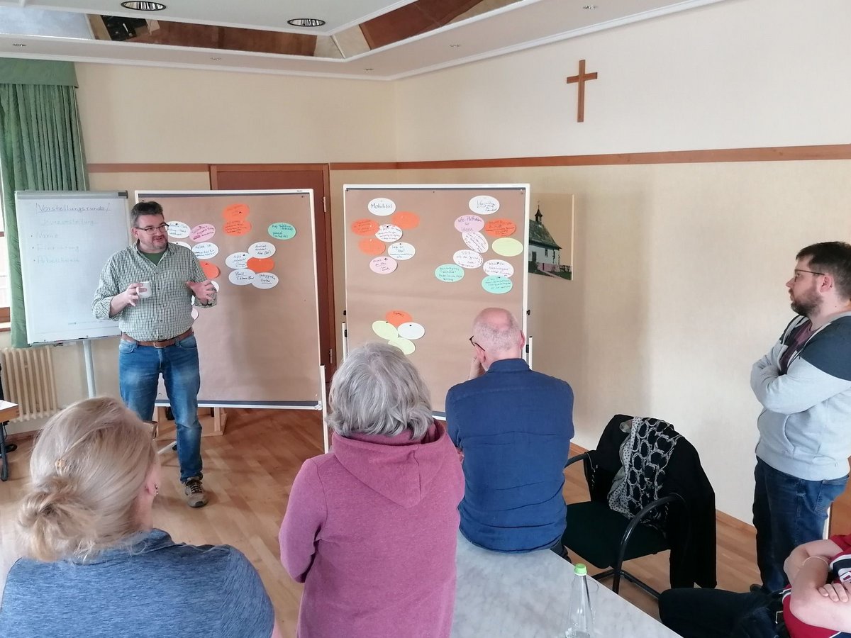 Ein Mann im karierten Hemd leitet einen Workshop für eine kleine Gruppe, mit Pinnwänden voller bunter Notizzettel.
