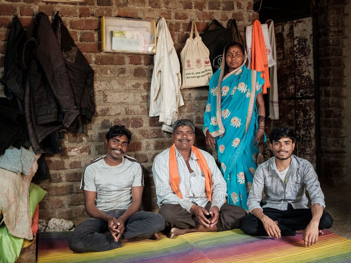 Eine Familie mit vier Personen (eine Frau, drei Männer) posiert in einem einfachen Raum mit Ziegelwand auf einer bunten Matte.
