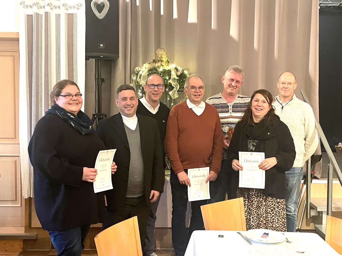 Acht lächelnde Personen, darunter ein Priester, halten Urkunden bei einer festlichen Veranstaltung.