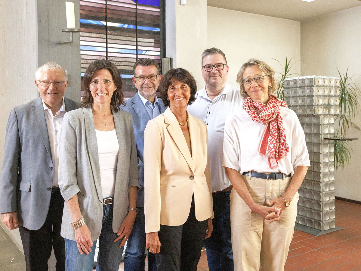 Sechs Personen, drei Frauen und drei Männer, lächeln in die Kamera. Sie stehen nebeneinander vor einem Buntglasfenster.