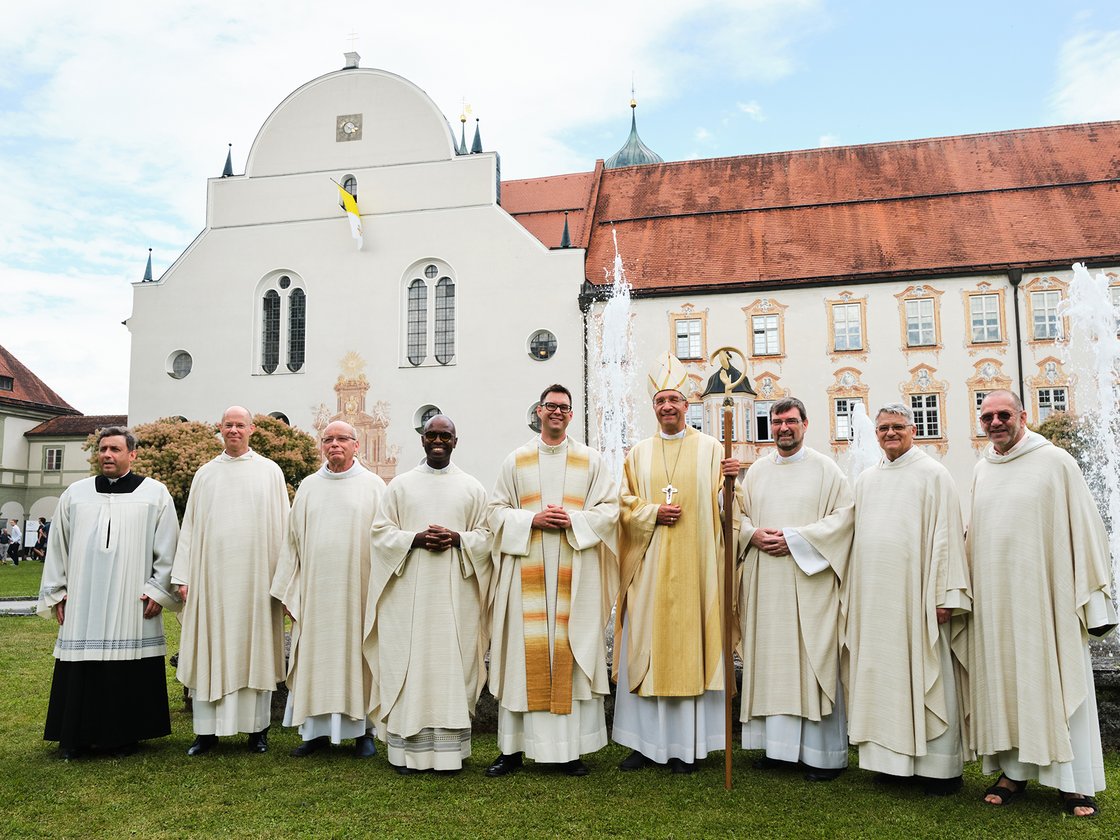 Gruppe von 9 Klerikern, darunter ein Bischof, auf Rasen vor einem weißen Klostergebäude mit Brunnen und Vatikanfahne.
