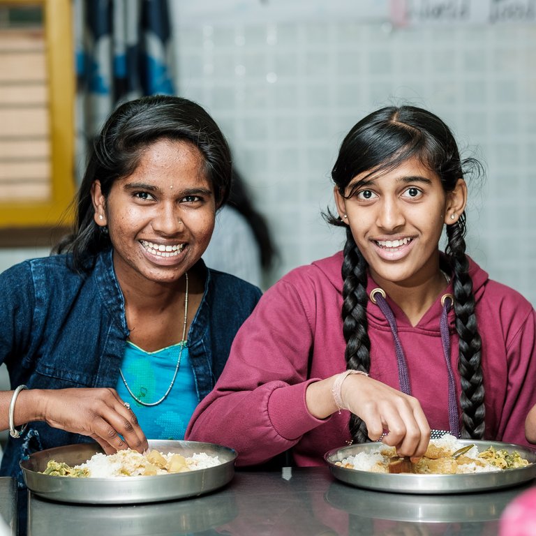Zwei lächelnde junge Frauen essen mit ihren Händen Reis und Curry von Metalltellern in einem Gemeinschaftsbereich.