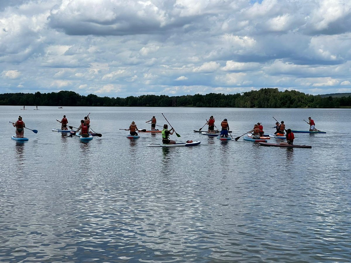 Eine Gruppe von Menschen beim Stand-Up-Paddling auf einem See vor bewaldetem Hintergrund unter bewölktem Himmel.