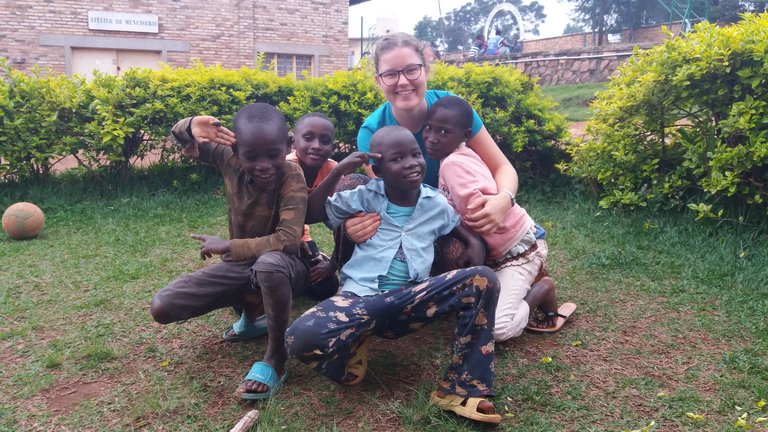 Eine lächelnde Frau mit Brille posiert mit fünf glücklichen Kindern draußen auf Gras, mit einem Gebäude und Büschen im Hintergrund.