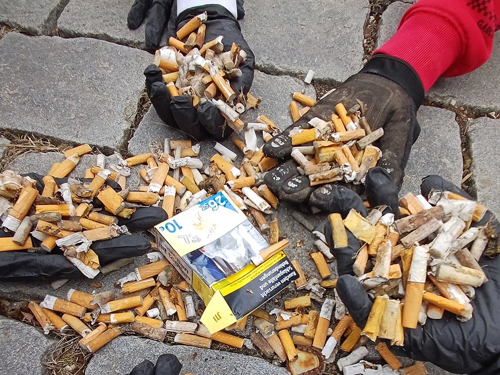 Behandschuhte Hände halten und sind umgeben von einem großen Haufen weggeworfener Zigarettenstummel und einer offenen Schachtel.