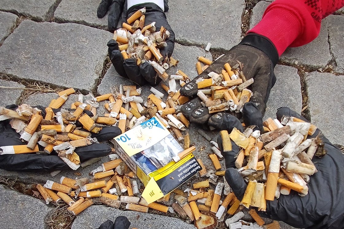 Behandschuhte Hände halten und sind umgeben von einem großen Haufen weggeworfener Zigarettenstummel und einer offenen Schachtel.