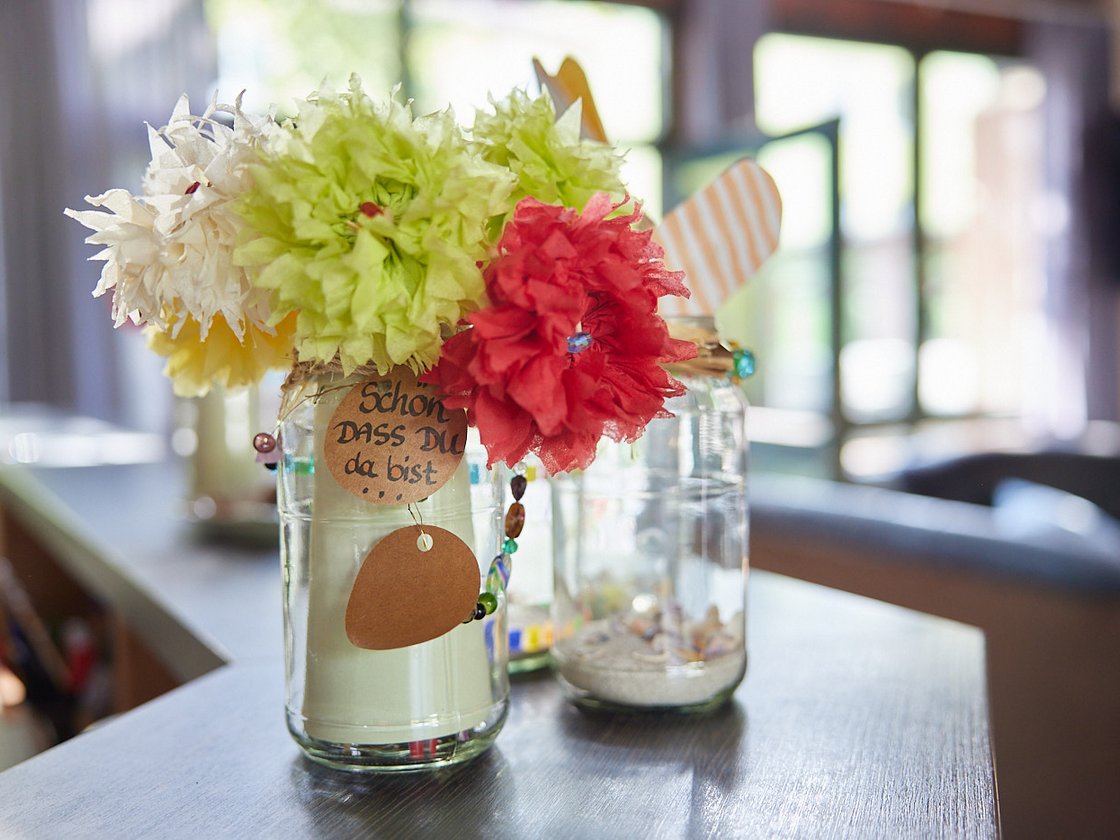 Drei dekorative Gläser mit bunten Papierblumen und Anhängern, eines mit der Aufschrift 'Schön, dass du da bist', auf einem Holztisch.