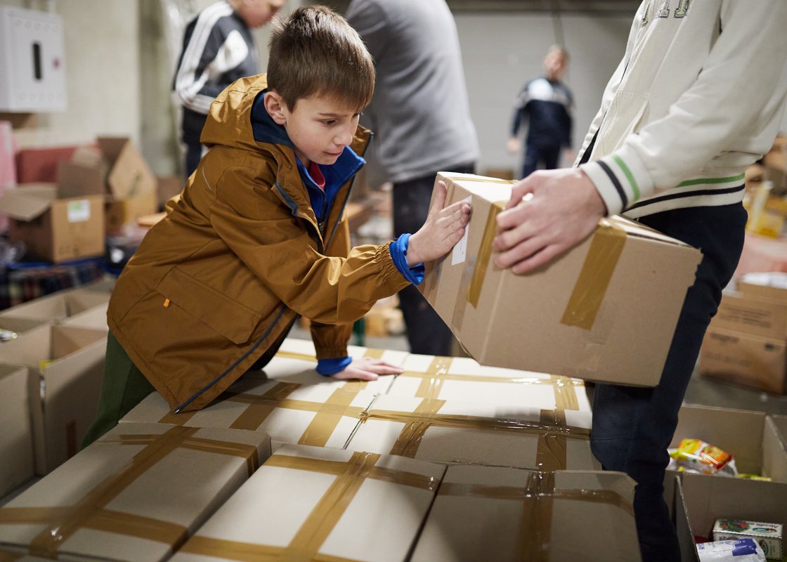 Ein Junge in brauner Jacke hilft einem Erwachsenen beim Stapeln von Hilfskartons in einem Lagerhaus.