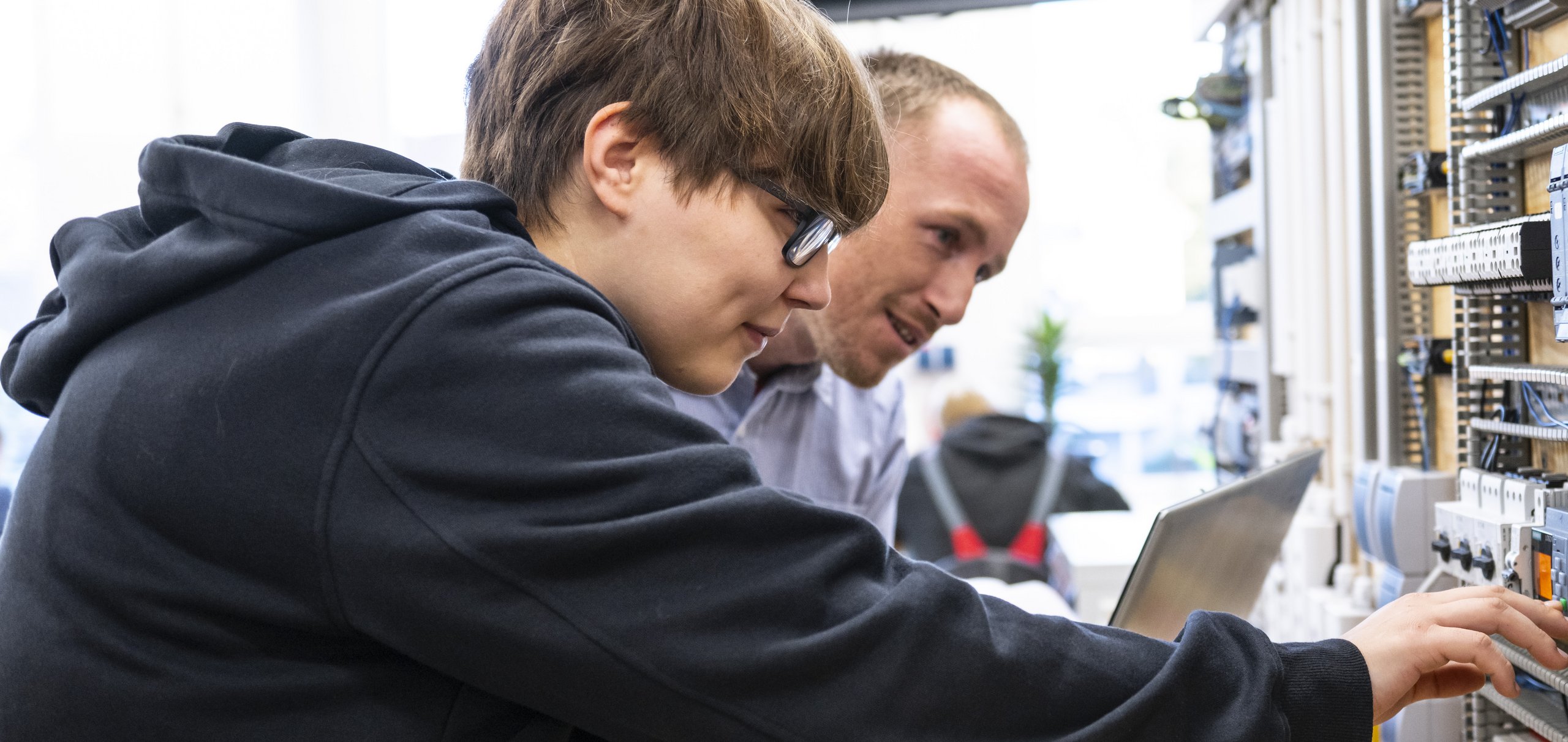 Zwei Techniker oder Studenten untersuchen eine elektrische Schalttafel mit Drähten und Modulen, einer zeigt auf ein Bauteil.