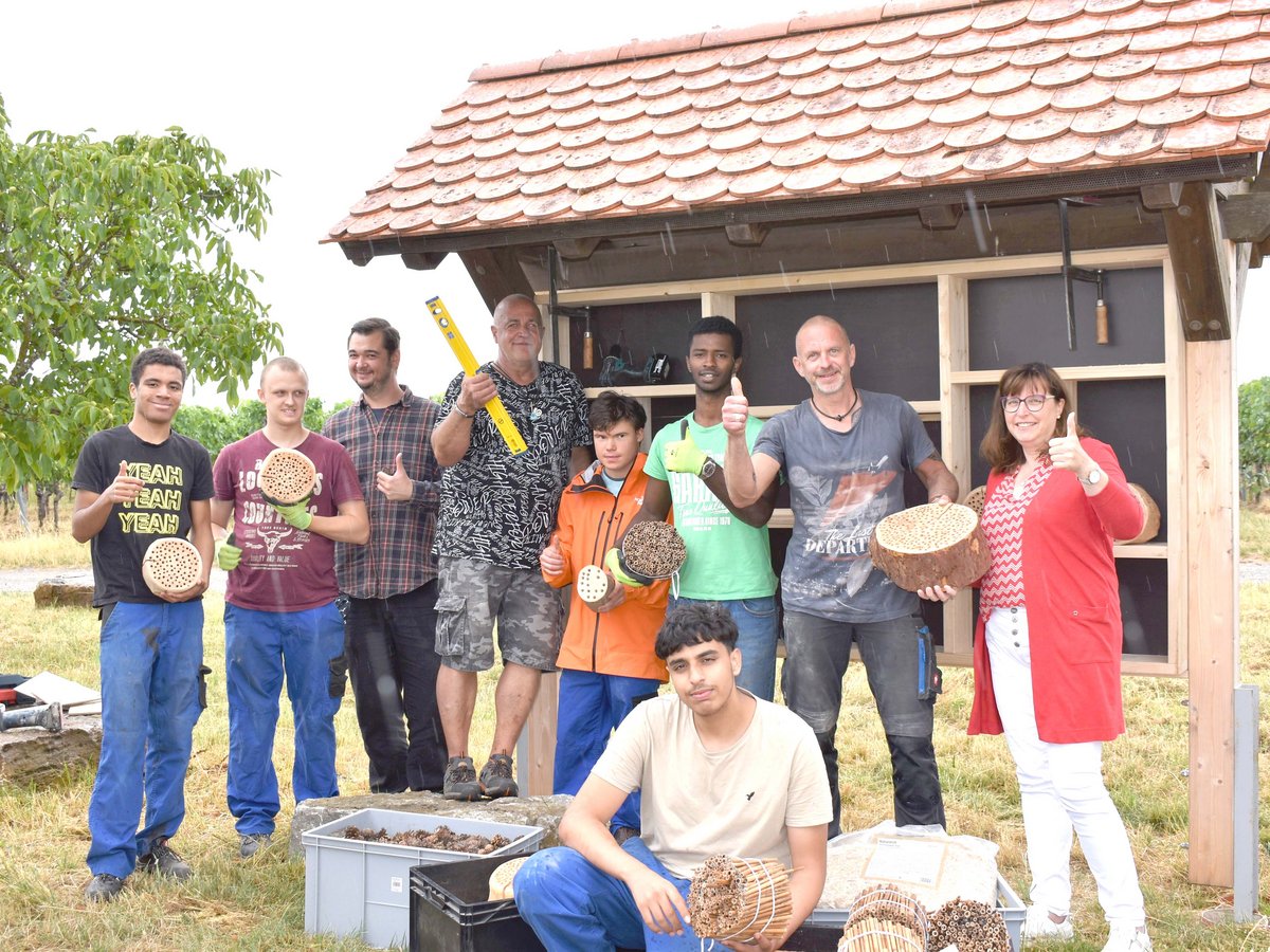 Eine Gruppe Menschen posiert stolz vor einem neuen Insektenhotel, hält Materialien wie gebohrte Holzscheiben und Bambusröhren und zeigt Daumen hoch, während es leicht regnet.