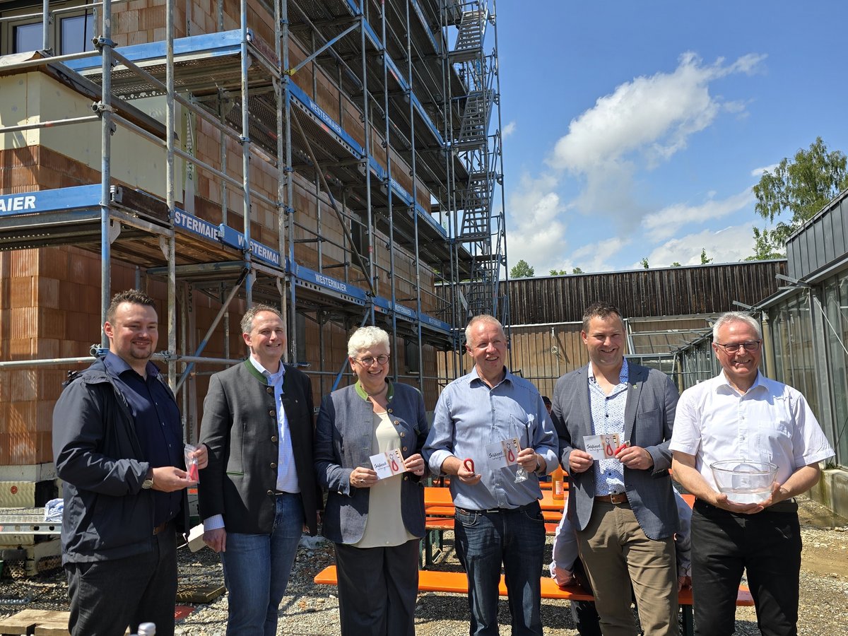 Sechs Personen lächeln bei einem Bau-Meilenstein vor einem eingerüsteten Gebäude, halten Schlüsselanhänger und Karten.