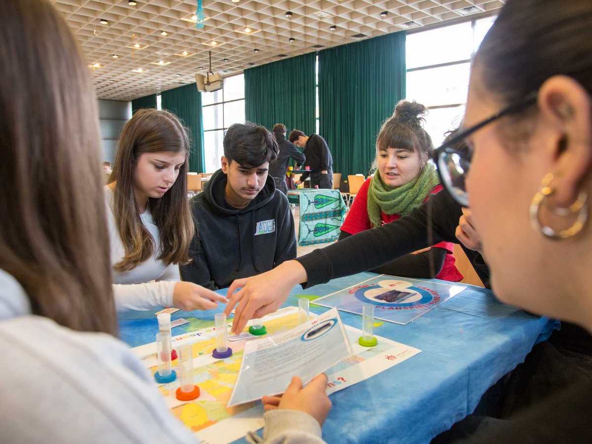 Schülerinnen und Schüler an einem Tisch spielen ein Lern-Brettspiel mit Reagenzgläsern und Karten.