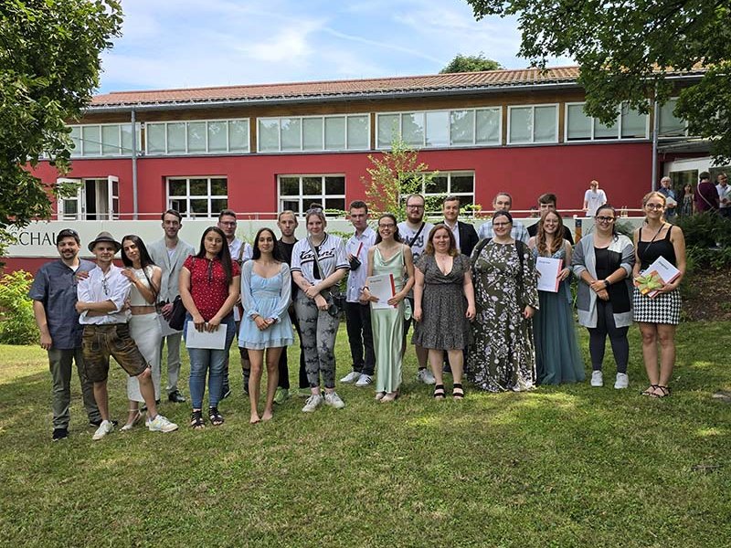 Eine vielfältige Gruppe Studenten und Absolventen posiert auf einem Rasen vor einem roten Gebäude, einige halten Diplome.